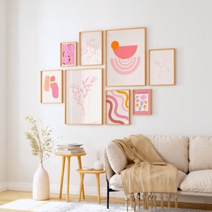 May include: A gallery wall with nine framed prints in various sizes and styles. The prints feature abstract shapes, floral designs, and line drawings in pink, orange, and yellow tones. The prints are arranged in a symmetrical pattern on a white wall.