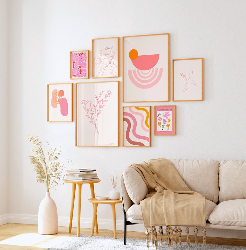 May include: A gallery wall with nine framed prints in various sizes and styles. The prints feature abstract shapes, floral designs, and line drawings in pink, orange, and yellow tones. The prints are arranged in a symmetrical pattern on a white wall.