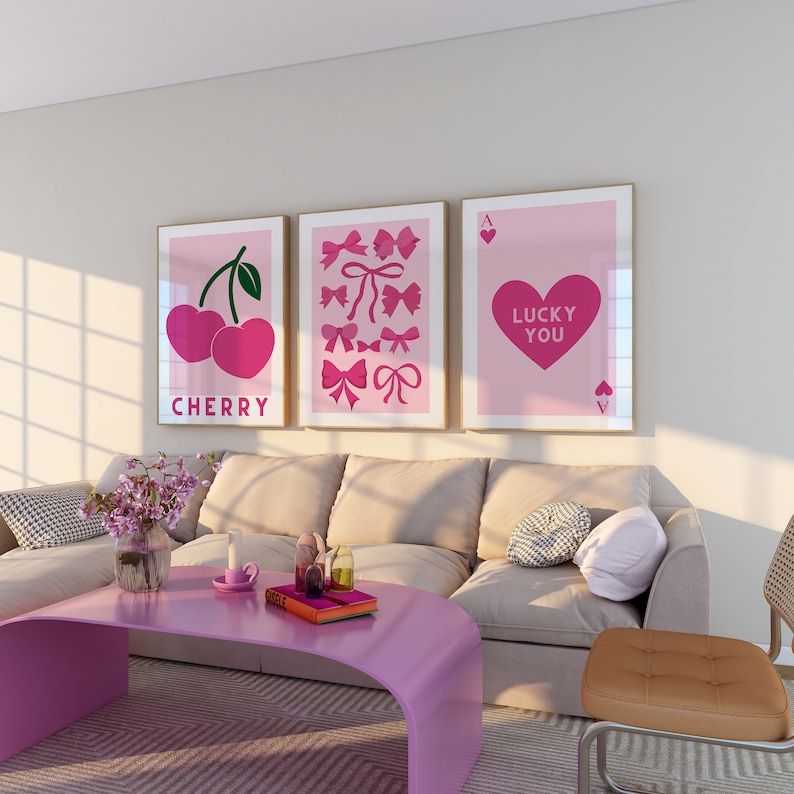 May include: Three pink framed prints on a white wall above a beige couch. The prints feature a cherry, a heart with the text "Lucky You", and a collection of pink bows.