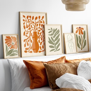 May include: Five framed prints featuring minimalist illustrations of plants and a human figure. The prints are in shades of orange, green, and white, and are arranged on a white wall above a bed with white and brown pillows.