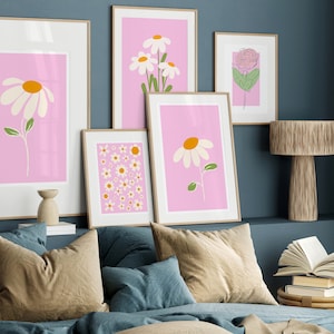 May include: A set of five framed prints featuring pink backgrounds with white and yellow daisy illustrations. The prints are arranged on a wall above a bed with brown and blue pillows.
