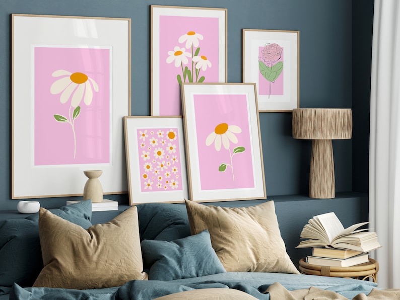 May include: A set of five framed prints featuring pink backgrounds with white and yellow daisy illustrations. The prints are arranged on a wall above a bed with brown and blue pillows.