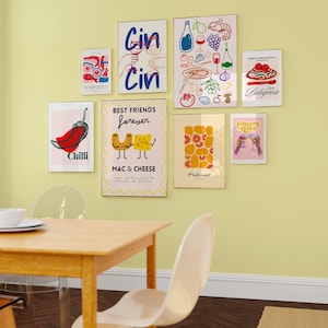May include: A collection of nine framed prints with various food and drink themes. The prints feature illustrations of chili peppers, mac and cheese, pizza, pasta, wine, and cocktails. The prints are arranged in a grid pattern on a light yellow wall above a wooden table and chairs.