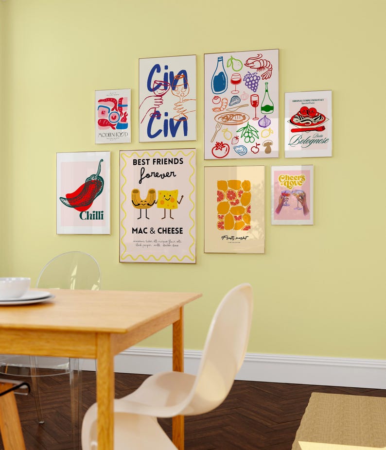 May include: A collection of nine framed prints with various food and drink themes. The prints feature illustrations of chili peppers, mac and cheese, pizza, pasta, wine, and cocktails. The prints are arranged in a grid pattern on a light yellow wall above a wooden table and chairs.