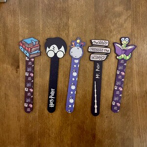 Könnte beinhalten: Fünf bunte Harry-Potter-Buchzeichen. Die Lesezeichen zeigen einen Stapel Bücher, Harry Potter mit Brille, eine Figur aus dem Kleinen Prinzen, einen Zauberstab und einen Wurm, der ein Buch liest.