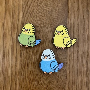 May include: Three wooden parakeet figurines, each with a different colour scheme. The first parakeet is green with black spots and a blue tail. The second parakeet is yellow with black spots. The third parakeet is blue with black spots and a purple tail. The parakeets are all facing forward with their beaks open.