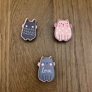May include: Three wooden cat figurines. The first cat is black with white eyes and a pink nose. The second cat is pink with white hearts and a white nose. The third cat is grey with pink cheeks and the word "love" written below it.