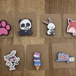 Puede incluir: Conjunto de diez pines de madera con varios personajes de dibujos animados. Los pines están sobre una superficie de madera marrón. Los personajes incluyen un panda, un conejo, un zorro, una huella de pata, un gato con gafas, un gato en un cono de helado y un conejo que sostiene cartas de juego.