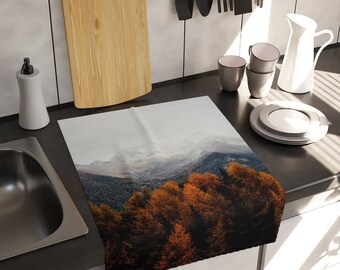 May include: A rectangular kitchen towel featuring a photographic print of a mountain range with snow-capped peaks above a dense forest of autumn trees with vibrant orange and dark brown foliage, displayed flat on a dark kitchen countertop.
