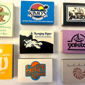 May include: A collection of vintage matchboxes featuring restaurant and hotel logos. The boxes display various designs, including the "Fish Tale" oyster bar, "KIMO'S" restaurant, and "Indian Wells Hotel". The matchboxes are rectangular and vary in color.