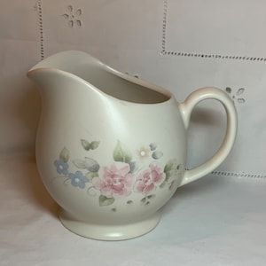 Könnte beinhalten: Ein weißer Keramik-Milchkännchen mit einem Blumendesign mit rosa und blauen Blumen. Der Milchkännchen hat einen abgerundeten Körper und einen Griff.