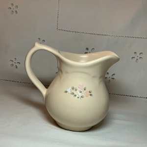 Könnte beinhalten: Ein cremefarbener Keramik-Milchkännchen mit einem Blumendesign in Rosa-, Orange- und Weißtönen. Der Milchkännchen hat einen runden Körper und einen gebogenen Griff.