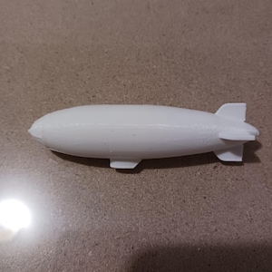 May include: A white, 3D-printed model of a zeppelin airship. The model features a rounded, elongated body with a fin and two small wings at the rear. The airship has two small wings on the bottom. The model is made of a solid, opaque white material.