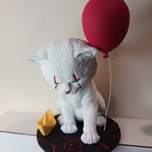May include: White cat figurine with red and green paint, holding a red balloon. The cat is sitting on a black base with a red and black pattern and the number "IT" written on it. A yellow paper boat is on the base.