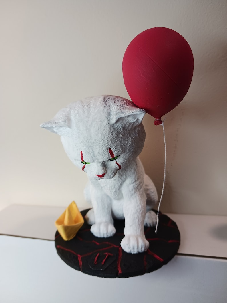 May include: White cat figurine with red and green paint, holding a red balloon. The cat is sitting on a black base with a red and black pattern and the number "IT" written on it. A yellow paper boat is on the base.