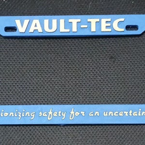 May include: Blue plastic license plate frame with the text "VAULT-TEC" in white letters and the Vault-Tec logo on either side. The bottom of the frame has the text "Revolutionizing safety for an uncertain future" in white letters.