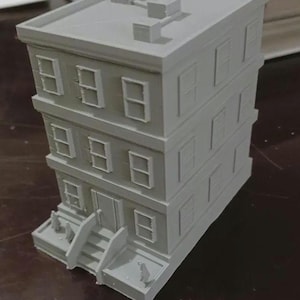 May include: A three-story, light gray model building with rectangular windows and a flat roof. The building has a front entrance with steps and small decorative elements. The model is likely for architectural or hobbyist use.