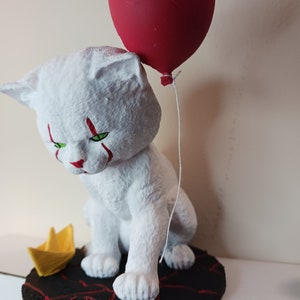May include: A white cat figurine with red clown makeup and green eyes, holding a red balloon. The cat is sitting on a black base with red cracks, and there is a yellow paper boat next to it.