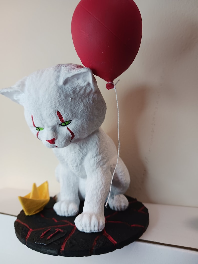 May include: A white cat figurine with red clown makeup and green eyes, holding a red balloon. The cat is sitting on a black base with red cracks, and there is a yellow paper boat next to it.