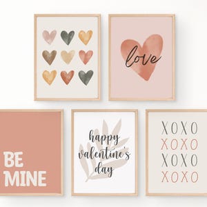 Może przedstawiać: Pięć oprawionych wydruków artystycznych z motywami walentynkowymi. Wydruki przedstawiają akwarelowe serca, słowo "love", frazę "Be Mine", "happy valentine's day" oraz litery "XOXO". Ramki są jasnobrązowe.