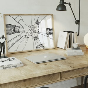 May include: A wooden desk with a framed black and white illustration of hands clasped together. A laptop, books, a lamp, a decorative vase, and a small sculpture are also on the desk. A moon-shaped lamp sits on a wooden stand.