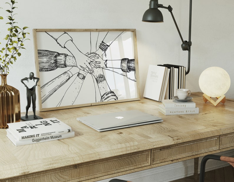 May include: A wooden desk with a framed black and white illustration of hands clasped together. A laptop, books, a lamp, a decorative vase, and a small sculpture are also on the desk. A moon-shaped lamp sits on a wooden stand.