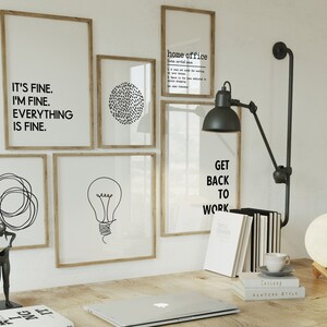 May include: A home office scene with framed art prints displaying motivational quotes and minimalist designs. A desk lamp, books, a laptop, and a moon lamp are visible. The artwork includes the phrases "IT'S FINE. I'M FINE. EVERYTHING IS FINE." and "GET BACK TO WORK."