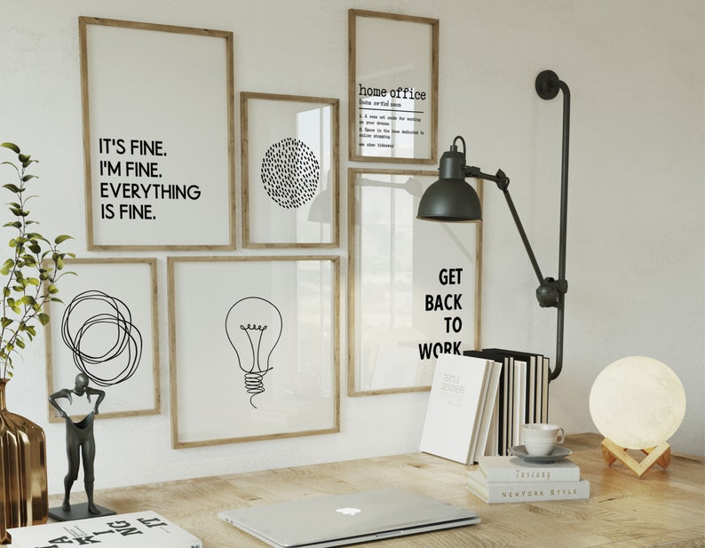 May include: A home office scene with framed art prints displaying motivational quotes and minimalist designs. A desk lamp, books, a laptop, and a moon lamp are visible. The artwork includes the phrases "IT'S FINE. I'M FINE. EVERYTHING IS FINE." and "GET BACK TO WORK."