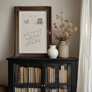 Pu&ograve; includere: Una stampa incorniciata di cartolina con il testo "quiet morning. empty tomb. full baskets." &egrave; appoggiata su un mobile nero pieno di libri. Un vaso bianco e un vaso rustico con fiori secchi sono anche sul mobile.