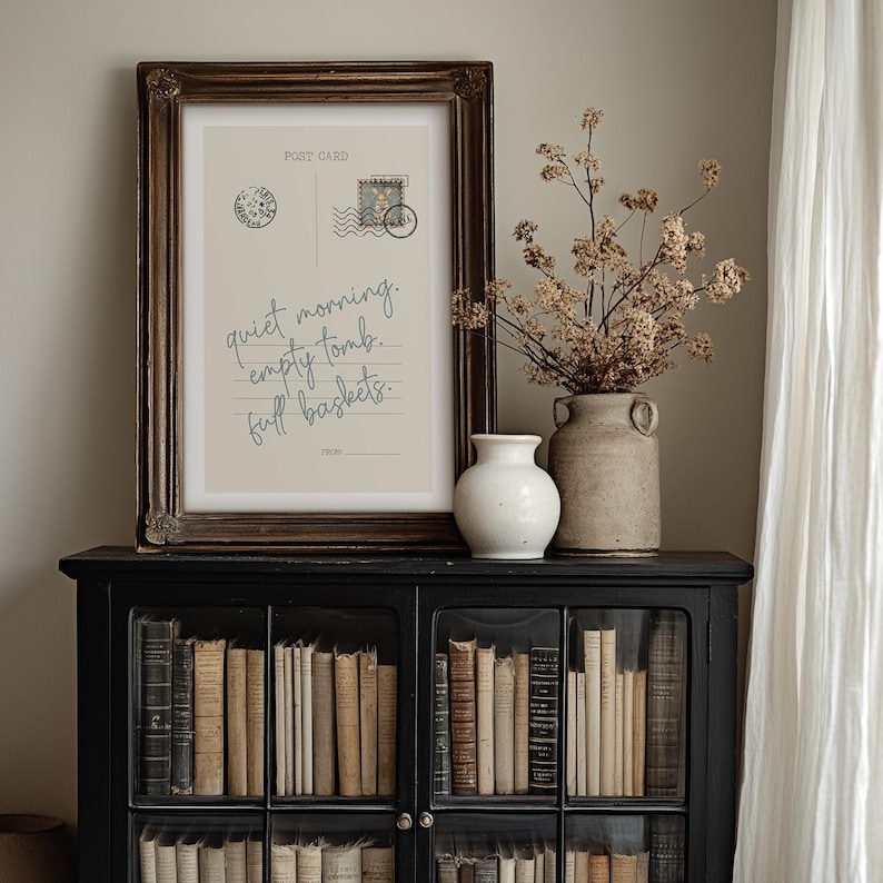 Pu&ograve; includere: Una stampa incorniciata di cartolina con il testo "quiet morning. empty tomb. full baskets." &egrave; appoggiata su un mobile nero pieno di libri. Un vaso bianco e un vaso rustico con fiori secchi sono anche sul mobile.