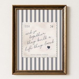 Puede incluir: Impresión enmarcada con una estética vintage. La obra de arte presenta un diseño de sello postal con el texto "and so together they built a life they loved" y un símbolo de corazón. El fondo tiene un patrón de rayas en azul y crema.