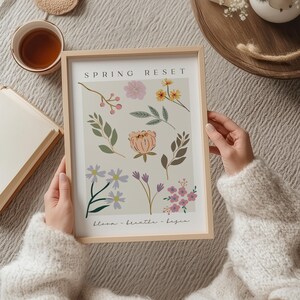 Pu&ograve; includere: Una stampa incorniciata con sfondo beige con illustrazioni floreali in rosa, giallo, viola e verde. Le parole "SPRING RESET" sono in alto, con "bloom - breathe - begin" in basso. La cornice &egrave; in legno chiaro.