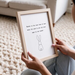 Pu&ograve; includere: Una stampa incorniciata con una cornice in legno chiaro. La stampa bianca presenta il testo "Easter is the only time of year when it's safe to put all your eggs in one basket" e un disegno a tratto di un coniglio.