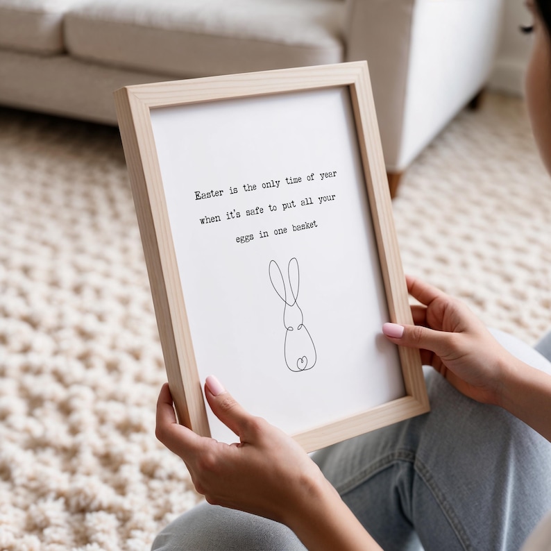 Pu&ograve; includere: Una stampa incorniciata con una cornice in legno chiaro. La stampa bianca presenta il testo "Easter is the only time of year when it's safe to put all your eggs in one basket" e un disegno a tratto di un coniglio.