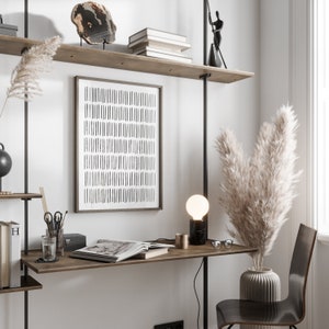 May include: A contemporary home office with a wooden desk and a framed abstract art print. A black marble lamp, decorative vase with pampas grass, and a wooden chair create a minimalist look. Shelves display books and decorative items.