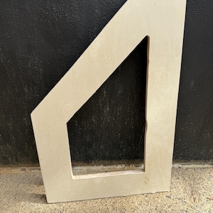 Puede incluir: Un marco de madera sin terminar de color claro, con un diseño angular. El marco tiene una abertura rectangular y está colocado sobre un fondo oscuro. La veta de la madera es visible, y el marco parece estar destinado a la personalización o al uso en un proyecto de artesanía.