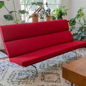 May include: A red three-cushioned sofa with a chrome frame sits on a white and gray rug in a living room.