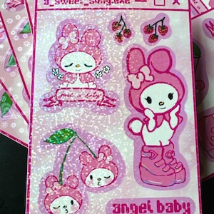 May include: Pink sticker sheet featuring cartoon characters with bunny ears, cherries, and the text "angel baby" and "a_sweet_song.exe". The stickers have a glittery finish and are in a kawaii style.