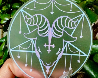 May include: A round sticker featuring a goat head with large horns, a cross, and a pentagram design. The sticker has a light green background with a light blue and purple design. Small stars are also included in the design.
