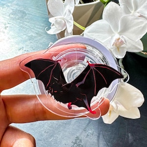 May include: A clear acrylic bat-shaped charm with a black body and red wing accents. The bat has its wings spread wide. The charm is round and has a clear border. The charm is a decorative item.