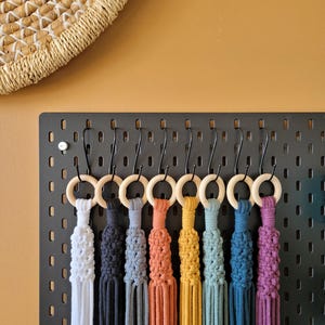 May include: A selection of colourful macrame plant hangers, each with a wooden ring, displayed on a black pegboard. The hangers are in shades of white, navy, grey, orange, yellow, teal, blue, and purple. A woven basket is visible in the background.