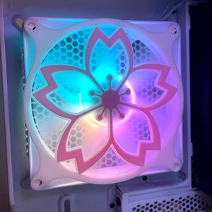 May include: A computer case fan with a white frame and a pink cherry blossom design. The fan blades are visible through the design, and the fan is lit with blue, pink, and yellow lights. The fan is mounted inside a computer case.