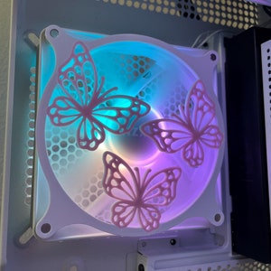 Puede incluir: Un ventilador de caja de ordenador blanco con tres recortes de mariposas rosas. El ventilador está iluminado con un degradado de luz azul y púrpura.
