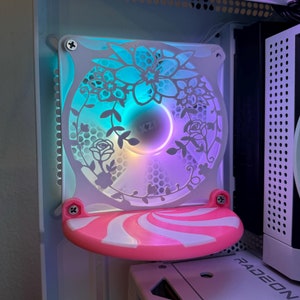 Op de afbeelding: Een witte computerbehuizingventilator met een roze en witte decoratieve afdekking. De ventilator heeft een bloemmotief en is verlicht met blauwe en paarse lampjes.