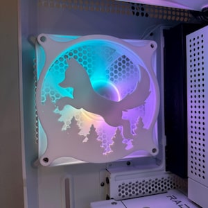 Könnte beinhalten: Ein weißer Computerlüfter mit einem ausgeschnittenen Design eines Fuchses, der durch einen Wald rennt. Der Lüfter wird mit einem Regenbogen aus Farben beleuchtet, darunter Blau, Grün, Lila und Gelb.