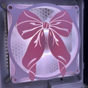 May include: A computer fan with a pink bow-shaped design. The fan is mounted inside a computer case, with a white and pink frame. The bow has a detailed cutout design, and the fan blades are visible behind it.