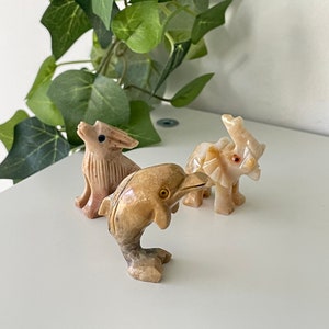 May include: Three small, hand-carved stone animal figurines. A light brown rabbit, a light brown dolphin, and a light brown elephant. The elephant has a red dot for an eye.