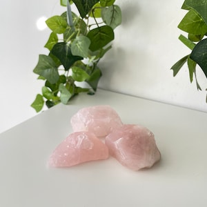 May include: Three pink rose quartz crystals on a white surface. The crystals are rough and have a natural, unpolished look.