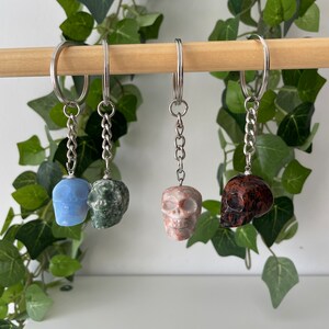 Puede incluir: Cuatro llaveros con forma de calavera hechos de diferentes tipos de piedra. Los llaveros est&aacute;n colgados de una varilla de madera. Las calaveras son azules, verdes, rosas y marrones.