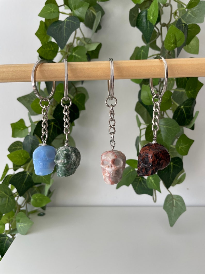 Puede incluir: Cuatro llaveros con forma de calavera hechos de diferentes tipos de piedra. Los llaveros est&aacute;n colgados de una varilla de madera. Las calaveras son azules, verdes, rosas y marrones.